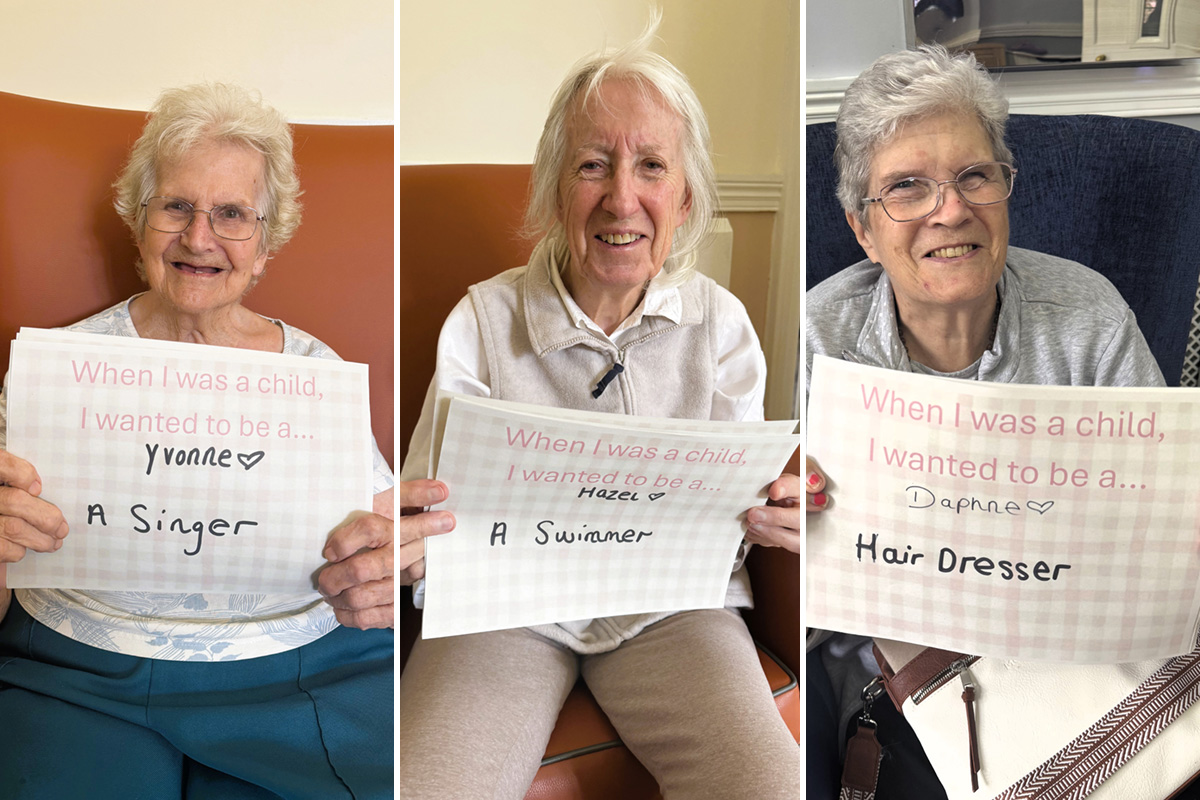 Three smiling female residents at The Old Downs Care Home holding handwritten signs that share their childhood dreams, including becoming a singer, a swimmer and a hairdresser.