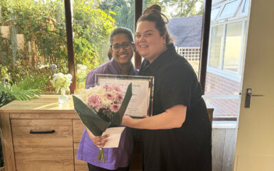 Two smiling team members at The Old Downs Residential Care Home, with one holding a bouquet of flowers and an Employee of the Month certificate.